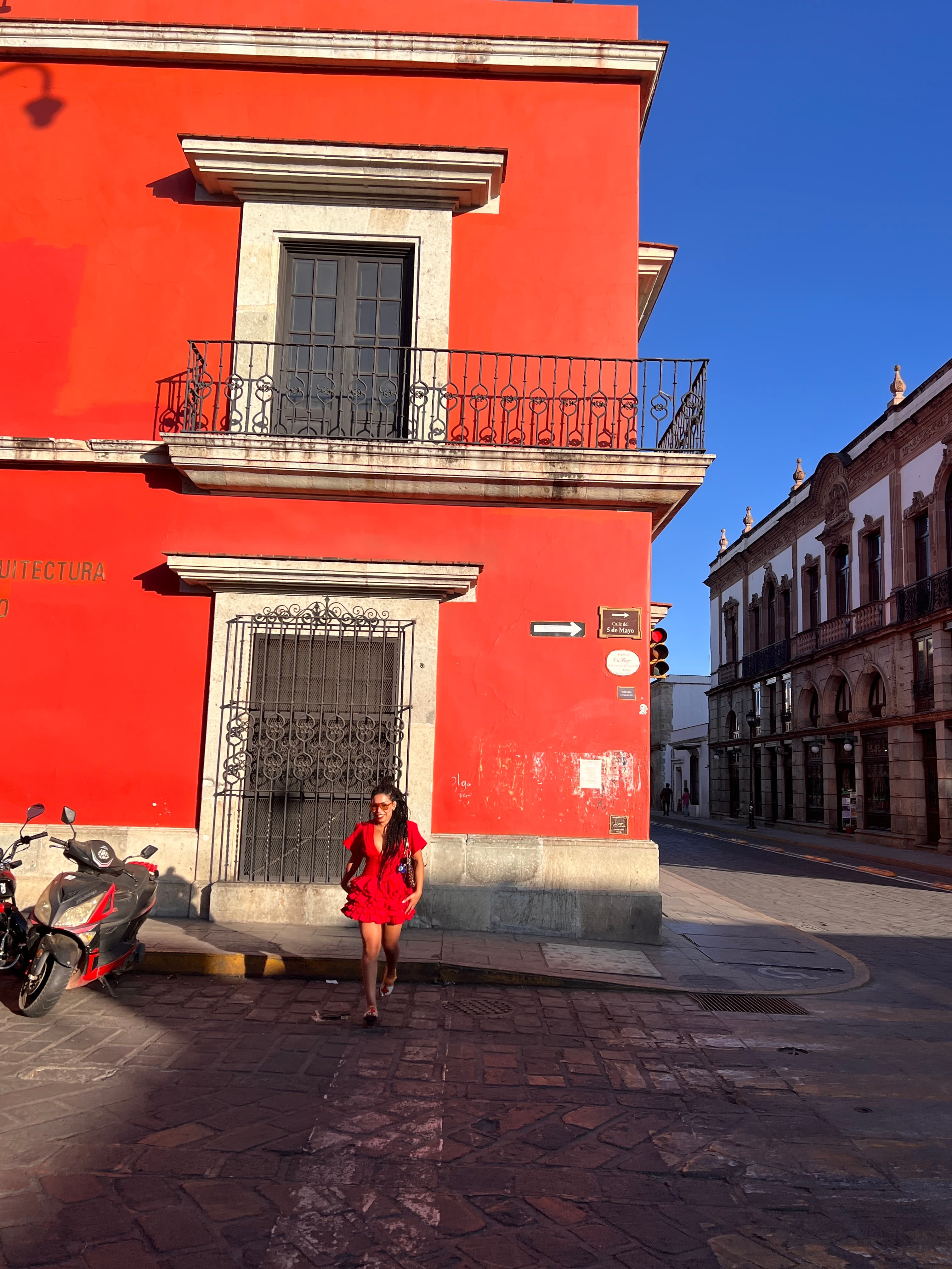 Sarah Moussem in a red dress crossing the road in Oaxaca, a red building behind her